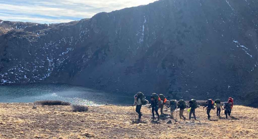 A group of people wearing backpacks hike in a line in front of a body of water in a mountainous landscape.